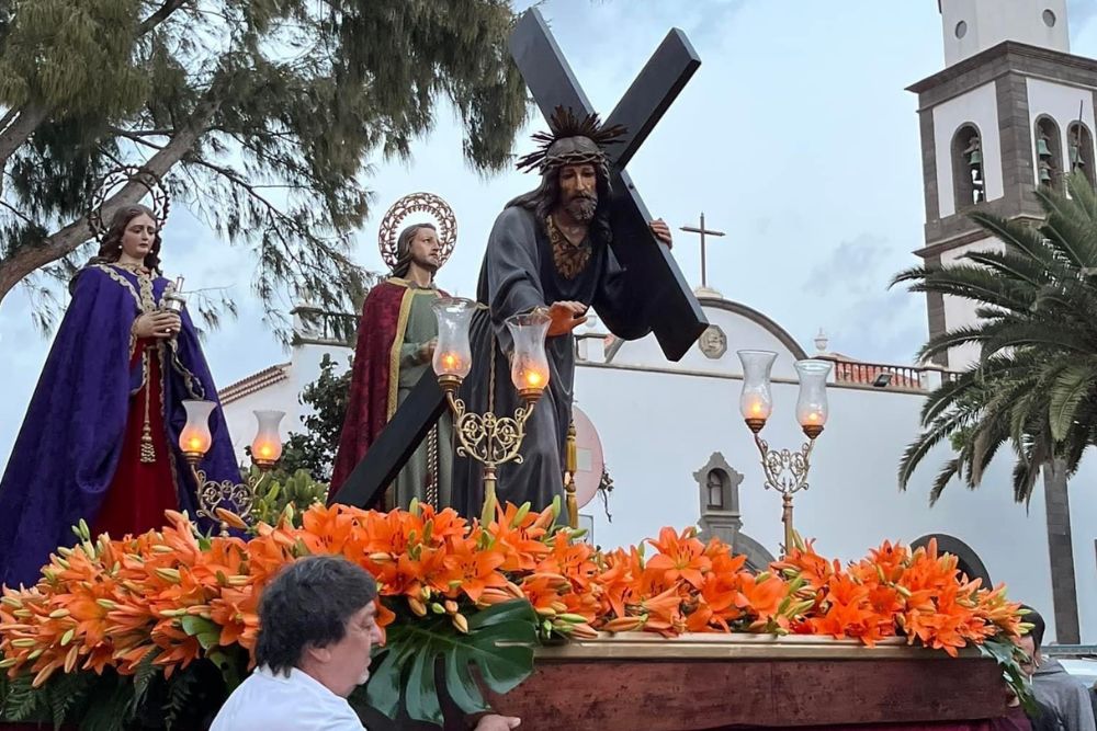 Paso procesional de Semana Santa en Lanzarote con Cristo portando la cruz, la Virgen y San Juan, decorado con lirios naranjas frente a una iglesia encalada