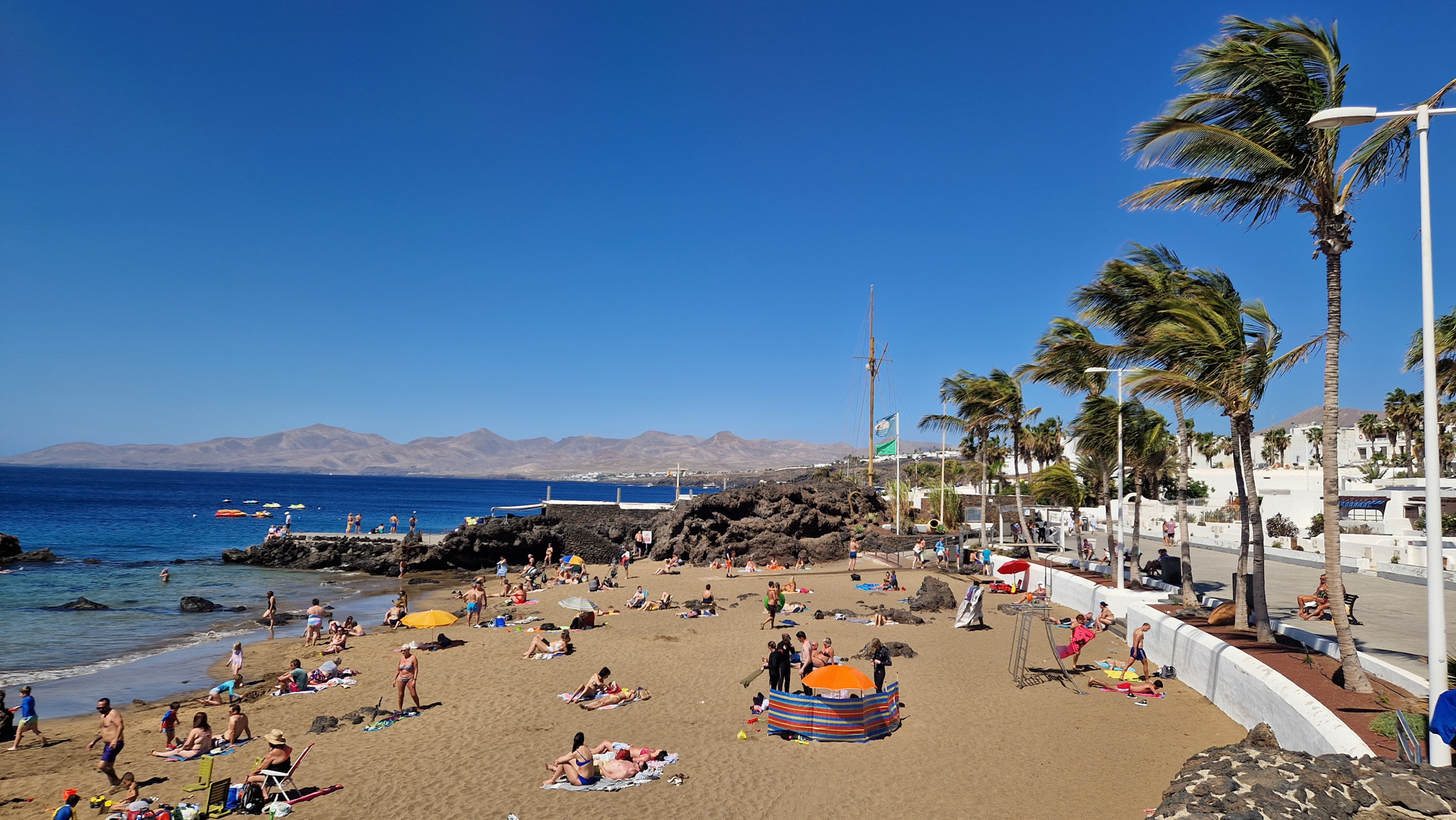 Playa de Lanzarote en junio con bañistas, palmeras y rocas volcánicas